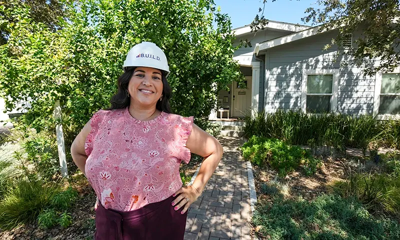 Alicia of BUILD wearing a branded hard hat standing in front of a house with greenery all around