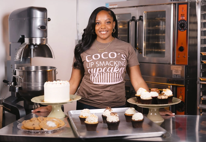 Nicole “Coco” Green of Coco's Lip Smacking Cupcakes standing in the kitchen in front of baking equipment and showcasing baked goods