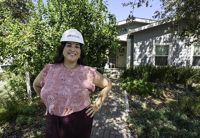Alicia of BUILD wearing a branded hard hat standing in front of a house with greenery all around